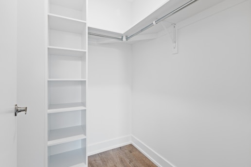 An empty walk-in closet with white walls, featuring built-in shelves on the left and a hanging rod extending along two walls. The floor is finished with a light brown wood texture, and there is a closed door with a metallic handle at the entrance.