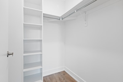 An empty walk-in closet with white walls, featuring built-in shelves on the left and a hanging rod extending along two walls. The floor is finished with a light brown wood texture, and there is a closed door with a metallic handle at the entrance.