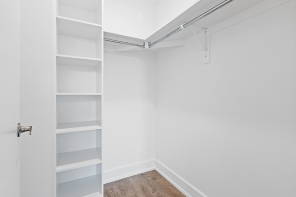 An empty walk-in closet with white walls, featuring built-in shelves on the left and a hanging rod extending along two walls. The floor is finished with a light brown wood texture, and there is a closed door with a metallic handle at the entrance.