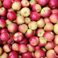 A large pile of fresh apples with a mix of red and green colors, tightly packed together. The apples vary slightly in size and have smooth, shiny skins.