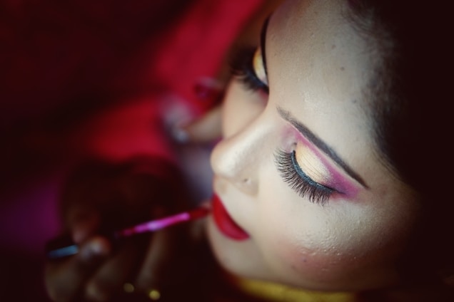 A close-up of a person having makeup applied to their face. The individual has vibrant, colorful eye makeup in shades of pink and gold, long eyelashes, and red lipstick. The person applying the makeup is holding a brush near the lips.