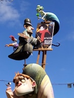 A whimsical sculpture featuring an artist character holding a palette and paintbrush, another character with exaggerated features holding a small bird, and a mermaid-like figure above them sipping from a glass. The sculpture is colorful and stands against a backdrop of a clear blue sky.