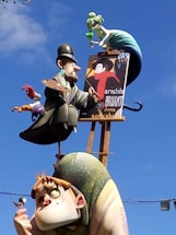 A whimsical sculpture featuring an artist character holding a palette and paintbrush, another character with exaggerated features holding a small bird, and a mermaid-like figure above them sipping from a glass. The sculpture is colorful and stands against a backdrop of a clear blue sky.