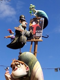 A whimsical sculpture featuring an artist character holding a palette and paintbrush, another character with exaggerated features holding a small bird, and a mermaid-like figure above them sipping from a glass. The sculpture is colorful and stands against a backdrop of a clear blue sky.