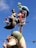 A whimsical sculpture featuring an artist character holding a palette and paintbrush, another character with exaggerated features holding a small bird, and a mermaid-like figure above them sipping from a glass. The sculpture is colorful and stands against a backdrop of a clear blue sky.