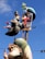A whimsical sculpture featuring an artist character holding a palette and paintbrush, another character with exaggerated features holding a small bird, and a mermaid-like figure above them sipping from a glass. The sculpture is colorful and stands against a backdrop of a clear blue sky.