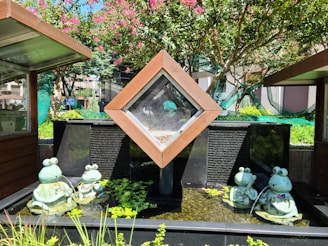 A water feature surrounded by green plants and trees, featuring frog sculptures in aquatic settings. Most frogs are depicted holding musical instruments or seated on leaves. The backdrop includes a wooden structure with a transparent panel and a textured stone wall.