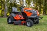 A bright orange riding lawn mower is parked on a well-manicured lawn. It features a large seat, steering wheel, and robust tires. Surrounding the mower are tall trees with green foliage, creating a serene outdoor setting.