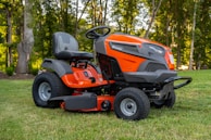 A bright orange riding lawn mower is parked on a well-manicured lawn. It features a large seat, steering wheel, and robust tires. Surrounding the mower are tall trees with green foliage, creating a serene outdoor setting.