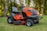 A bright orange riding lawn mower is parked on a well-manicured lawn. It features a large seat, steering wheel, and robust tires. Surrounding the mower are tall trees with green foliage, creating a serene outdoor setting.