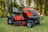 A bright orange riding lawn mower is parked on a well-manicured lawn. It features a large seat, steering wheel, and robust tires. Surrounding the mower are tall trees with green foliage, creating a serene outdoor setting.