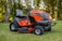 A bright orange riding lawn mower is parked on a well-manicured lawn. It features a large seat, steering wheel, and robust tires. Surrounding the mower are tall trees with green foliage, creating a serene outdoor setting.