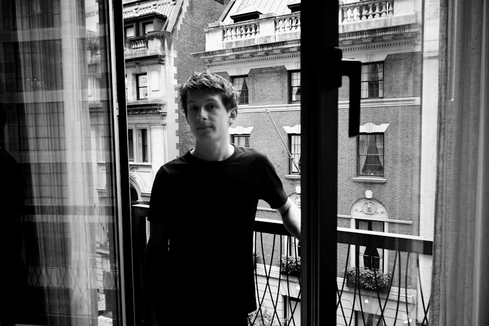 Foto en escala de grises de un hombre con camiseta negra de cuello redondo de pie cerca de una ventana de vidrio