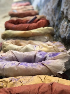 A series of fabric bundles covered in various colored powders are lined up along a textured surface. The colors include shades of purple, yellow, red, and brown, suggesting a vivid and earthy palette often used in artistic or crafting activities.