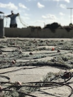 A fishing net is sprawled across the ground with orange and red floats attached to it. In the background, a person is standing and holding part of the net. The sky is bright with scattered clouds and there is a muted, coastal atmosphere.