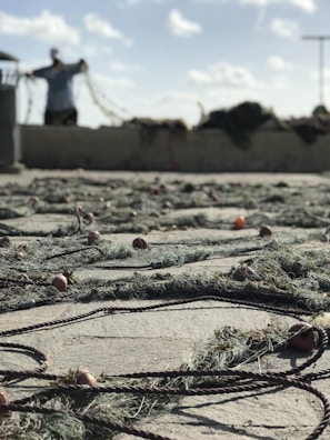 A fishing net is sprawled across the ground with orange and red floats attached to it. In the background, a person is standing and holding part of the net. The sky is bright with scattered clouds and there is a muted, coastal atmosphere.