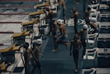 Young swimmers wearing swim caps and swimsuits are preparing to compete at an indoor swimming pool. They are gathered near starting blocks on the pool deck, appearing to be in different stages of warm-up exercises. The environment has a competitive and focused atmosphere, with pool lanes visible in the water.