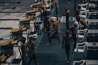 Young swimmers wearing swim caps and swimsuits are preparing to compete at an indoor swimming pool. They are gathered near starting blocks on the pool deck, appearing to be in different stages of warm-up exercises. The environment has a competitive and focused atmosphere, with pool lanes visible in the water.