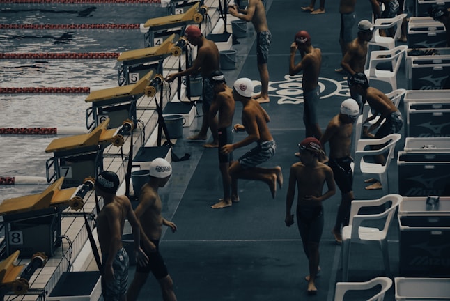 Young swimmers wearing swim caps and swimsuits are preparing to compete at an indoor swimming pool. They are gathered near starting blocks on the pool deck, appearing to be in different stages of warm-up exercises. The environment has a competitive and focused atmosphere, with pool lanes visible in the water.