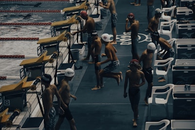 Young swimmers wearing swim caps and swimsuits are preparing to compete at an indoor swimming pool. They are gathered near starting blocks on the pool deck, appearing to be in different stages of warm-up exercises. The environment has a competitive and focused atmosphere, with pool lanes visible in the water.