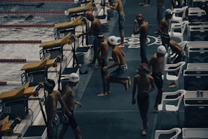 Young swimmers wearing swim caps and swimsuits are preparing to compete at an indoor swimming pool. They are gathered near starting blocks on the pool deck, appearing to be in different stages of warm-up exercises. The environment has a competitive and focused atmosphere, with pool lanes visible in the water.