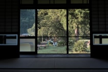 A traditional Japanese room with tatami flooring opens to a serene view of a lush garden. Large sliding glass doors reveal the vibrant greenery outside, including tall trees and well-maintained shrubs. Soft natural light filters through, creating a peaceful and harmonious atmosphere.