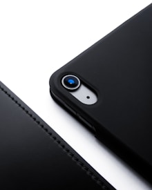 A close-up view of a smartphone with a black case, focusing on the camera lens. The phone is placed on a white surface, creating a stark contrast. The case has a smooth texture and precise cutouts for the camera and flash.