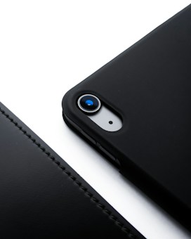 A close-up view of a smartphone with a black case, focusing on the camera lens. The phone is placed on a white surface, creating a stark contrast. The case has a smooth texture and precise cutouts for the camera and flash.