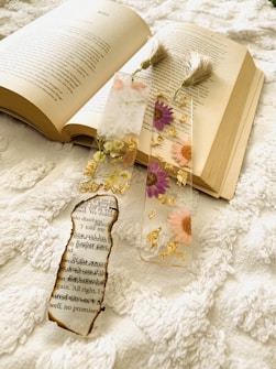 An open book lies on a textured white fabric, accompanied by three decorative bookmarks. One bookmark features a piece of burnt paper with text, while the other two contain colorful pressed flowers and gold accents. Both have tassels attached at the top.