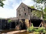 A rustic wooden mill sits by a flowing stream, featuring a large waterwheel attached to the side of the building. The mill is constructed with dark brown, weathered wooden planks and rests on a stone foundation. Surrounding the mill are lush green trees and shrubbery that add a natural frame to the scene. The water flows gently over stones next to the mill, creating a serene atmosphere.