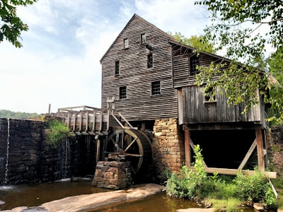A rustic wooden mill sits by a flowing stream, featuring a large waterwheel attached to the side of the building. The mill is constructed with dark brown, weathered wooden planks and rests on a stone foundation. Surrounding the mill are lush green trees and shrubbery that add a natural frame to the scene. The water flows gently over stones next to the mill, creating a serene atmosphere.