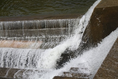 Water cascades over a series of concrete steps in a structured man-made waterfall. The rapid flow creates white frothy waves as it descends, with water glistening under natural light. The structured edges suggest an urban or industrial design.