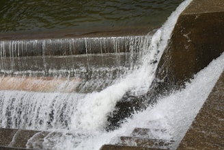 Water cascades over a series of concrete steps in a structured man-made waterfall. The rapid flow creates white frothy waves as it descends, with water glistening under natural light. The structured edges suggest an urban or industrial design.