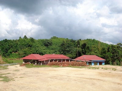 A rural scene features two houses with red roofs set against a backdrop of dense, green trees under a mostly cloudy sky. Piles of bricks are stacked in front of the buildings, suggesting ongoing or recent construction work. The area around the houses is mostly open, with patches of grass on the sandy ground.
