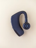 A black Bluetooth earpiece with a sleek design is placed on a light beige background. The earpiece has an ergonomic shape with a circular speaker grill featuring a concentric pattern.