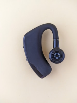 A black Bluetooth earpiece with a sleek design is placed on a light beige background. The earpiece has an ergonomic shape with a circular speaker grill featuring a concentric pattern.