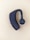 A black Bluetooth earpiece with a sleek design is placed on a light beige background. The earpiece has an ergonomic shape with a circular speaker grill featuring a concentric pattern.
