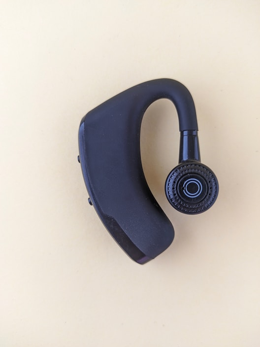A black Bluetooth earpiece with a sleek design is placed on a light beige background. The earpiece has an ergonomic shape with a circular speaker grill featuring a concentric pattern.