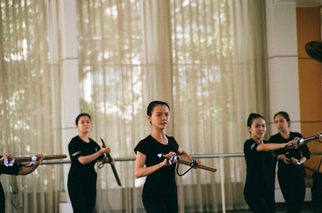A group of women practicing martial arts in a well-lit room, each holding a wooden or bamboo training weapon. They are wearing matching black attire and appear focused and coordinated, suggesting a structured training session.