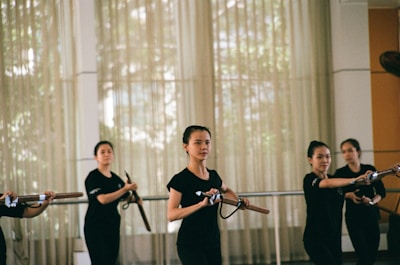 A group of women practicing martial arts in a well-lit room, each holding a wooden or bamboo training weapon. They are wearing matching black attire and appear focused and coordinated, suggesting a structured training session.