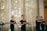 A group of women practicing martial arts in a well-lit room, each holding a wooden or bamboo training weapon. They are wearing matching black attire and appear focused and coordinated, suggesting a structured training session.