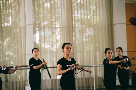 A group of women practicing martial arts in a well-lit room, each holding a wooden or bamboo training weapon. They are wearing matching black attire and appear focused and coordinated, suggesting a structured training session.