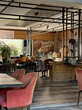 A cozy interior space with warm, inviting decor featuring wooden furniture and red velvet chairs. The walls have a mix of wooden panels and brick, and there's an artistic mural on one wall. Large windows let in natural light, enhancing the relaxed atmosphere.