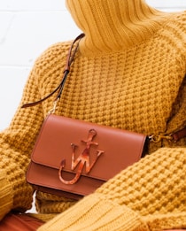 A person wearing a textured mustard yellow turtleneck sweater is holding a brown leather handbag with a unique emblem design on the front. The setting is minimalistic, with a focus on the fashion items.