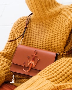 A person wearing a textured mustard yellow turtleneck sweater is holding a brown leather handbag with a unique emblem design on the front. The setting is minimalistic, with a focus on the fashion items.