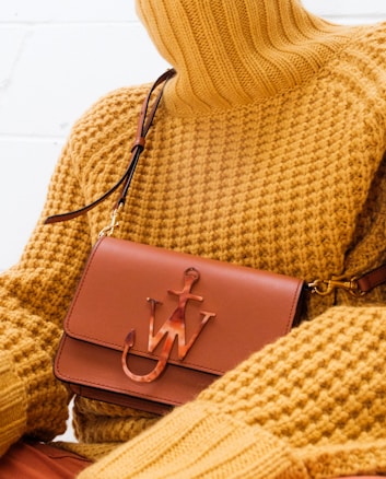 A person wearing a textured mustard yellow turtleneck sweater is holding a brown leather handbag with a unique emblem design on the front. The setting is minimalistic, with a focus on the fashion items.