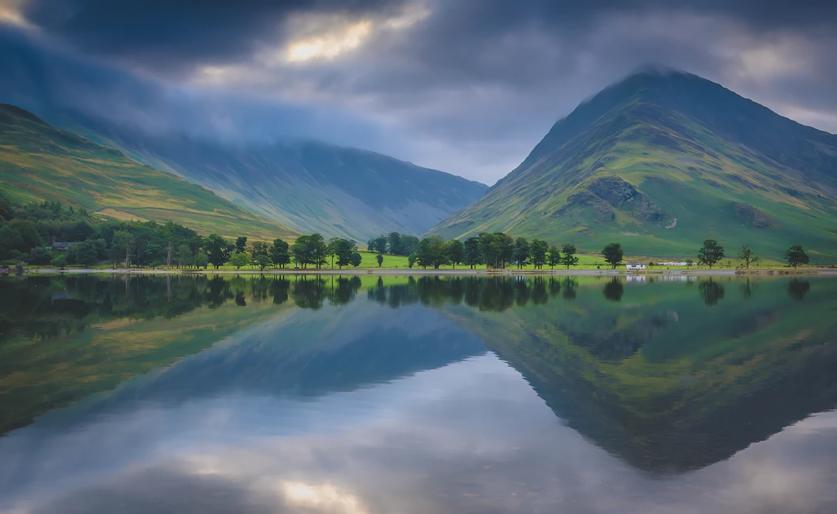 Green fells reflected in still water at Buttermere, with a line of trees along the far shore