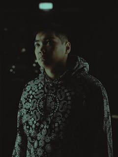 A person in a dark environment, wearing a hooded sweatshirt with a distinctive paisley pattern. The lighting casts shadows on their face, creating a dramatic effect.