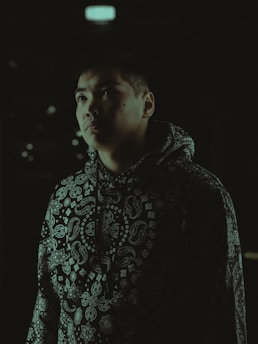 A person in a dark environment, wearing a hooded sweatshirt with a distinctive paisley pattern. The lighting casts shadows on their face, creating a dramatic effect.