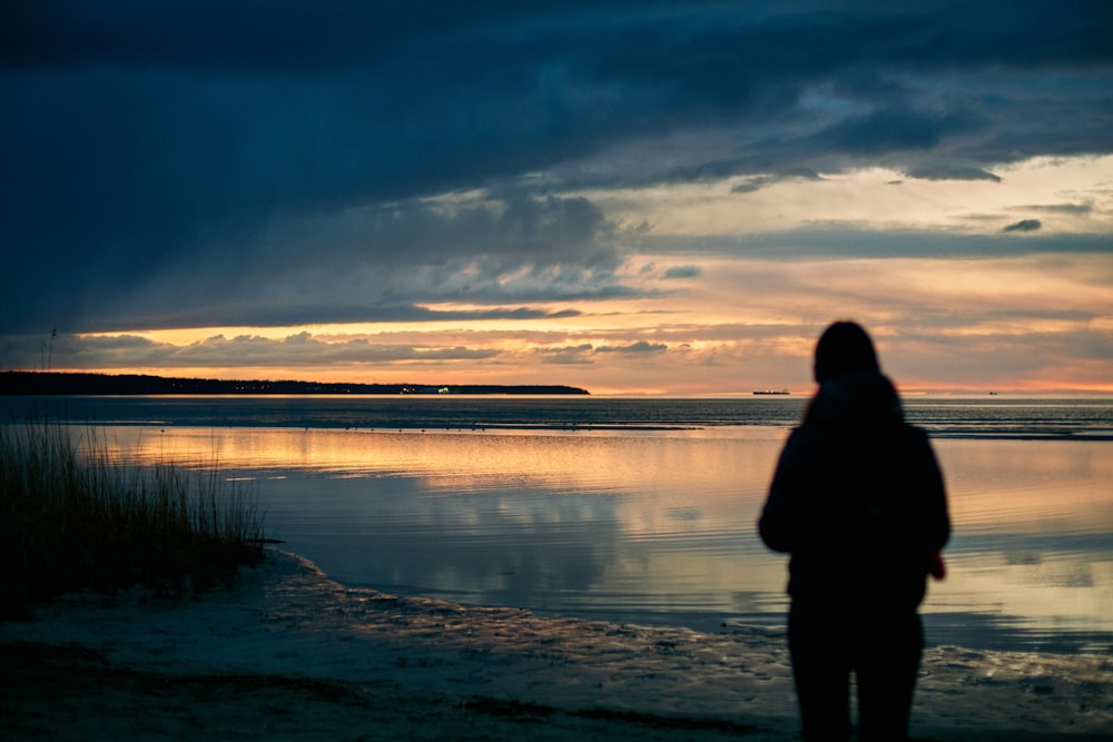 silhouette of person standing near body of water under cloudy sky during daytime